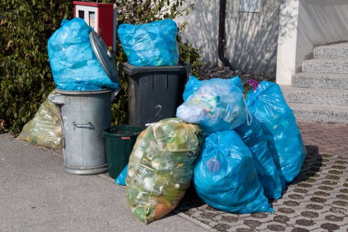 A collection of overflowing rubbish bags and waste bins situated on a paved urban sidewalk area, near a building with steps. The rubbish bags are mainly blue and transparent, filled with various types of household waste, including plastic bottles and paper. Some bags are tied at the top, while others appear loosely packed. There are several bins visible, including a grey metal bin on the left, a dark grey plastic bin in the center, and a small green bin positioned in front of the larger containers. The background includes a low wall or fence with greenery and some shrubs behind it. The scene suggests a typical waste collection spot that may require rubbish removal services in the area, such as those provided by Islington House Clearance. The overall setting indicates a busy residential or commercial street, with the waste temporarily accumulated before collection or removal. The lighting is natural, suggesting daytime conditions, and the area looks clean aside from the gathered rubbish bags.