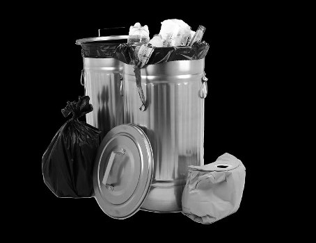 A large metallic rubbish bin with a fitted lid, filled with mixed waste including visible crumpled paper and plastic packaging, situated on a dark surface against a black background. Next to the bin are a tied black rubbish bag and a smaller crumpled paper bag, indicating a waste collection set-up typical of domestic or property clearance in London. The trash is organized around the bin, with the lid resting on the ground, suggesting recent disposal activity. The overall scene reflects professional rubbish removal services offered by Islington House Clearance, emphasizing efficient waste management for residential properties within the local postcode area. The metallic finish of the bin shows slight reflections under lighting, and the waste items are casual and unstacked, accurately conveying typical rubbish materials encountered during clearance or disposal tasks.