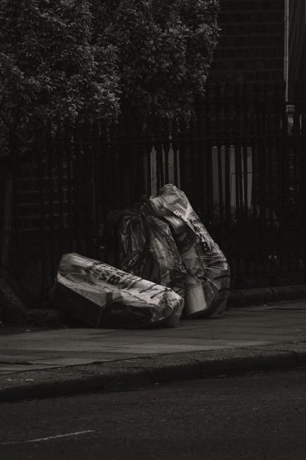 A group of large black plastic rubbish bags filled with discarded waste are placed on a sidewalk next to a curb. The bags are crumpled and tied at the top, with some displaying printed text or logos. 