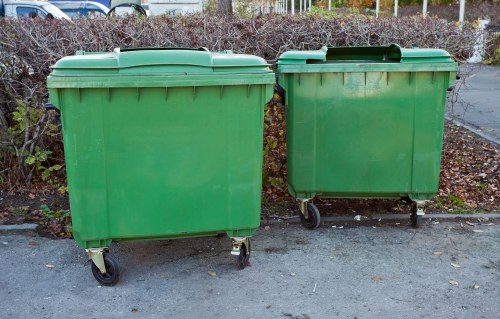 Two large green plastic wheelie bins with black wheels and lids are positioned side by side on a paved surface in an outdoor area. They are situated in front of a background consisting of a hedge with brown and purple foliage, indicating a late autumn or early winter setting in an urban or residential environment. The bins are clean and appear to be used for rubbish collection, with slight scuff marks on their surfaces. The setting suggests a typical rubbish disposal point near a property, possibly in Islington or a nearby area, consistent with local waste management practices in London. The image is clear, with the bins centrally located and occupying most of the frame, emphasizing their size and practical use in waste collection and clearance services, such as those provided by Islington House Clearance.
